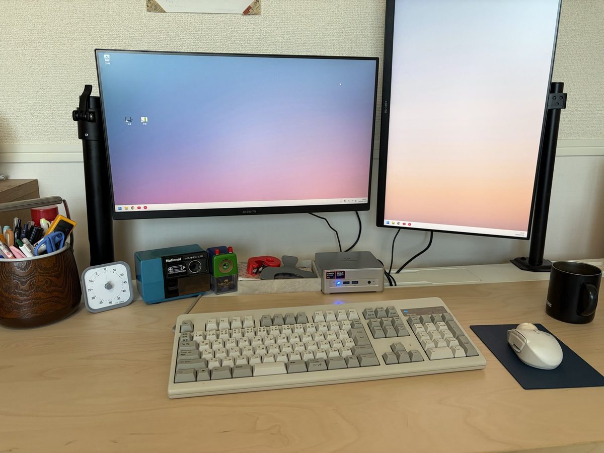 新しいPCを置いた机。横長のモニターと縦向きのモニターが並び、手前に白いキーボードとマウス、モニターの下に手のひらサイズのミニPC本体が置いてある